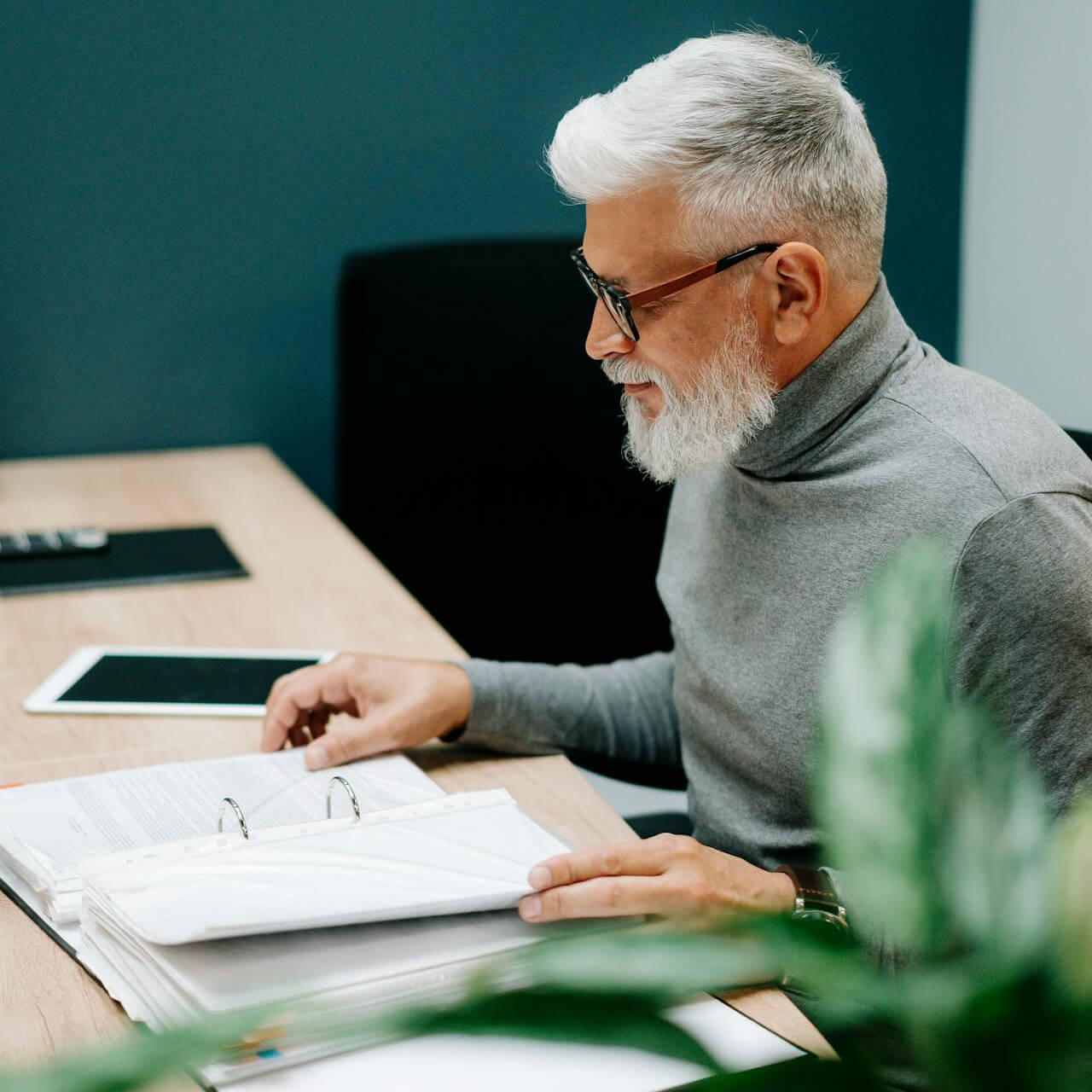 Ein älterer Mann mit grauem Haar und Brille sitzt an einem Tisch und betrachtet ein Dokument in einem Ordner. Neben ihm liegt ein Tablet.