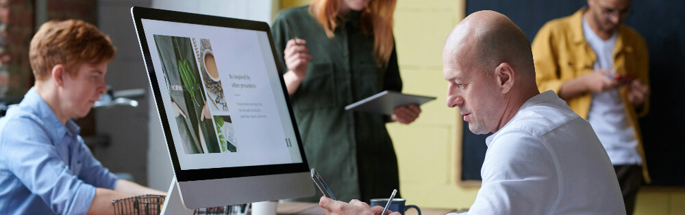 Eine Gruppe von Personen arbeitet gemeinsam in einem modernen Büro. Im Vordergrund sitzt ein Mann vor einem Computerbildschirm, der eine Website zeigt. Im Hintergrund stehen zwei Personen, die ein Tablet und ein Smartphone benutzen. Ein weiterer Mann sitzt seitlich im Bild an einem Tisch.