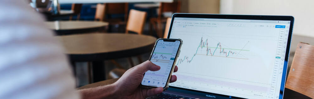 Altersvorsorge mit Fonds Eine Person sitzt an einem Tisch mit einem Laptop und hält ein Smartphone in der Hand. Der Laptop zeigt ein Diagramm mit Kursmustern und Trendlinien.